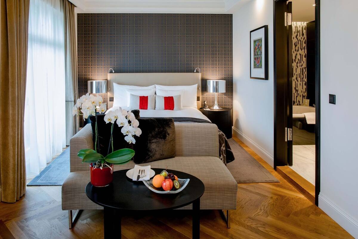 A modern hotel room with a bed, bedside tables, a sofa, a dining table with fruit, and a view of the bathroom.