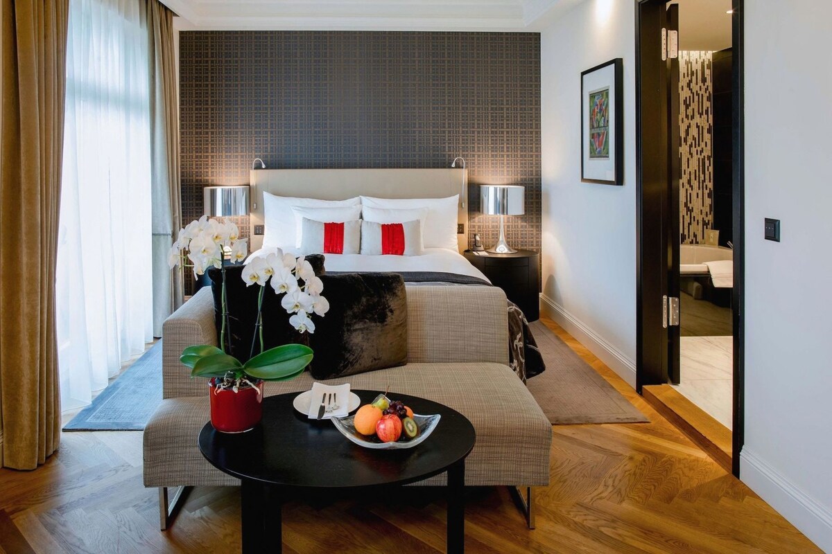 A hotel room with a bed, bedside tables, a desk with a fruit bowl, a potted orchid, and a view of the bathroom.
