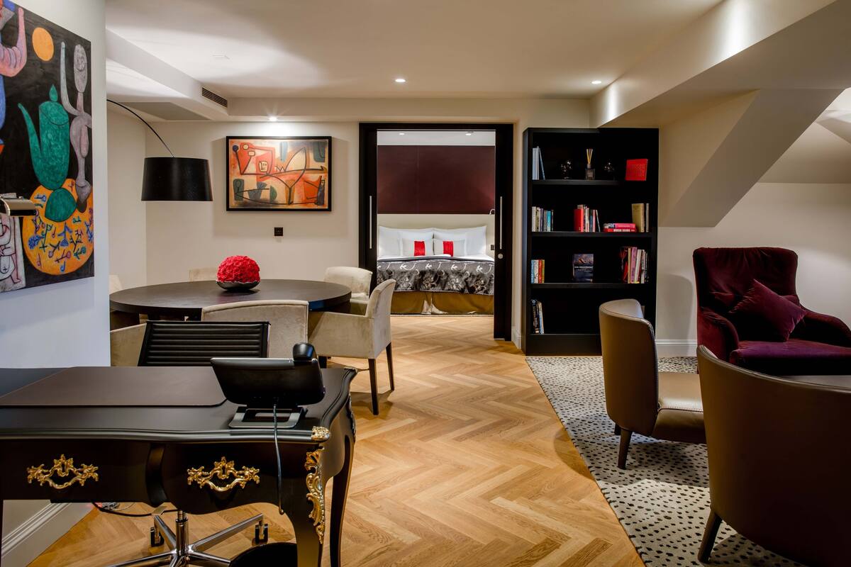 A modern living room with a wooden herringbone floor, a round dining table with chairs, a red vase, a black bookshelf, and a red armchair.