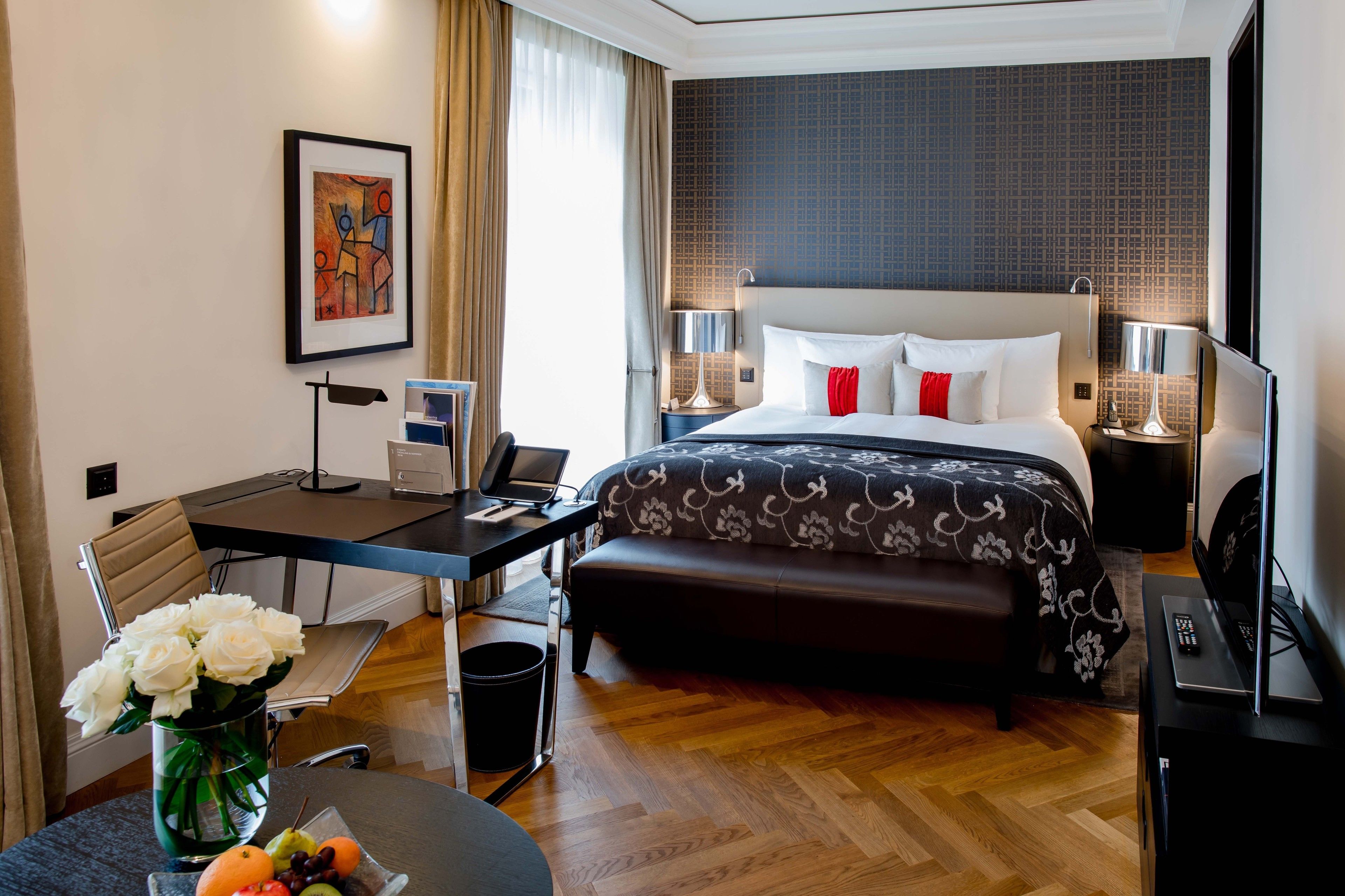A hotel room with a bed, desk, chair, and a small table with a vase of flowers.