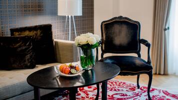 A room with a round black table, a vase with white flowers, and a tray with fruits. There is a black upholstered chair and a patterned rug.