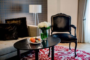 A room with a round black table, a vase with white flowers, and a tray with fruits. There is a black upholstered chair and a patterned rug.