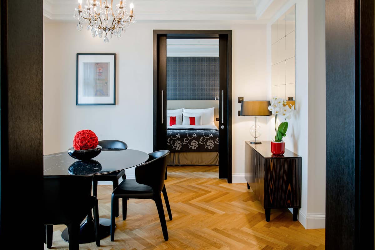 A modern hotel room with a wooden floor, a dining area with a black table and chairs, a bed with a dark headboard, a bedside table with a lamp, and a framed picture on the wall.