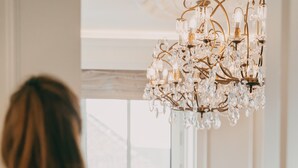 A person opening a door to a room with a large window, a chandelier, and a vase of flowers on the windowsill.