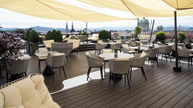 Terraza al aire libre con mesas redondas y sillas de mimbre, con vista a un paisaje urbano que incluye edificios destacados y una estructura con cúpula.