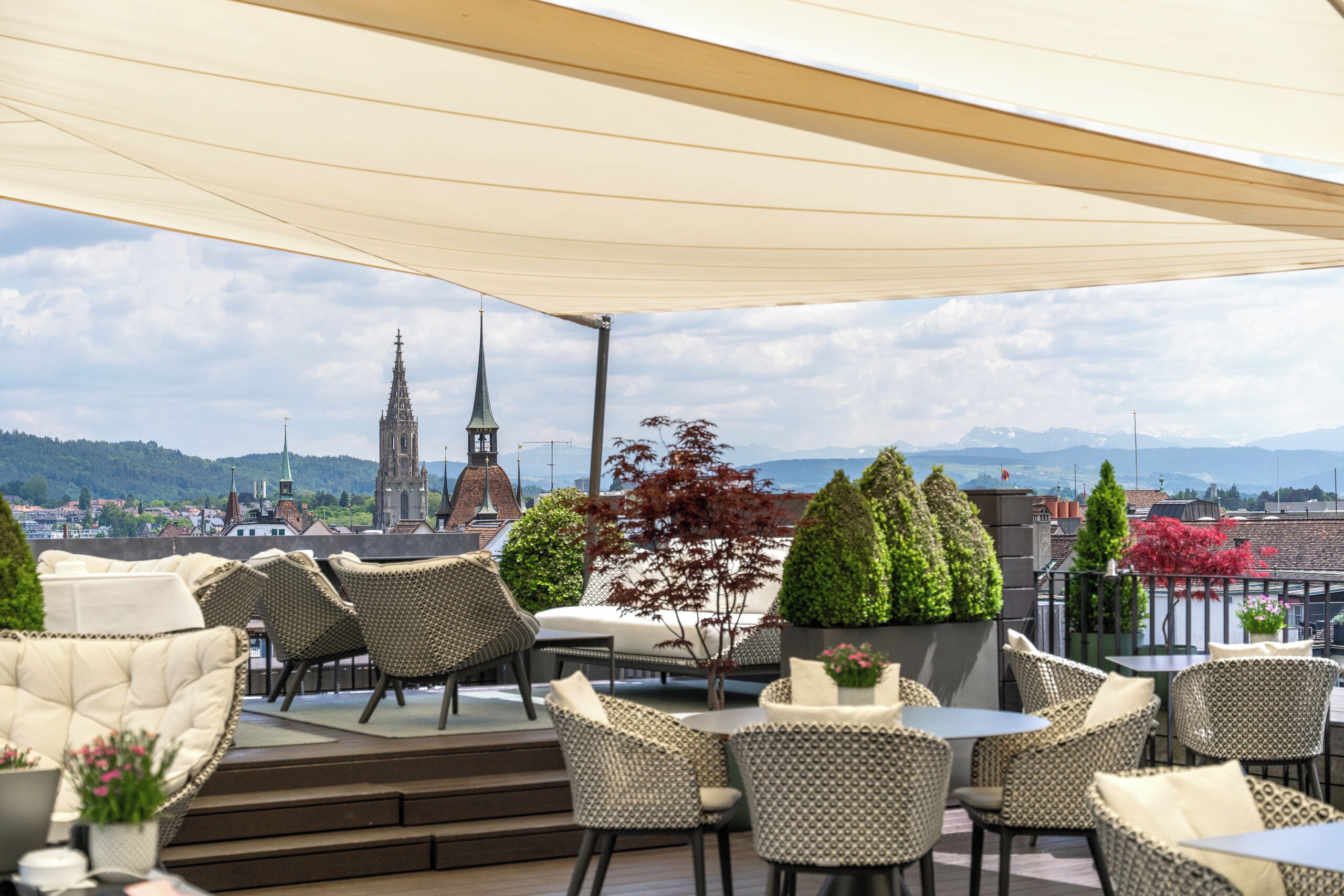 An outdoor seating area with wicker furniture, a view of a cityscape including a church tower, and a mountain range in the distance.