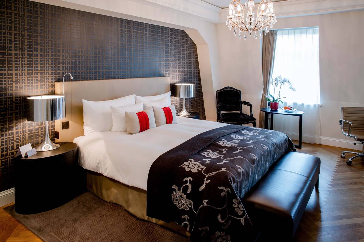 A modern hotel room with a large bed, two bedside tables with lamps, a bench, a chair, a small table with a plant, and a chandelier.