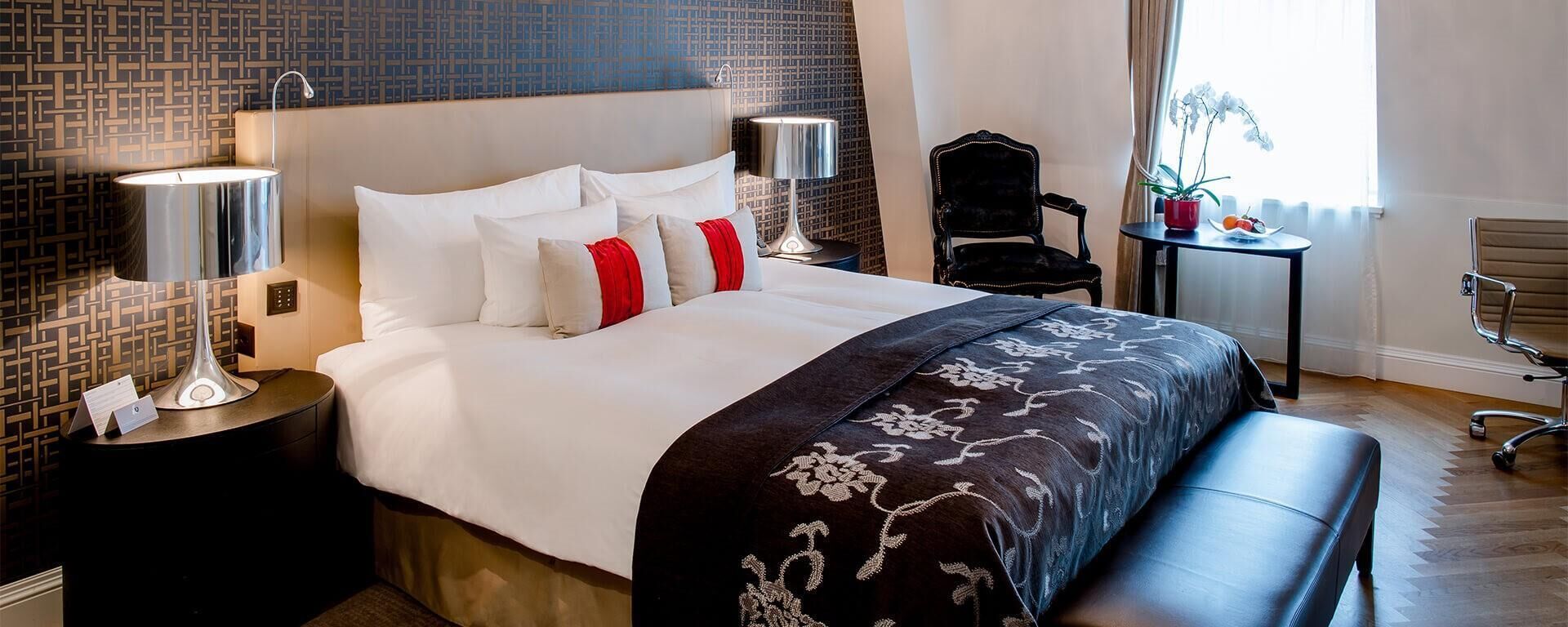 A hotel room with a large bed, two bedside tables with lamps, a chair, a small table with a plant, and a window with curtains.