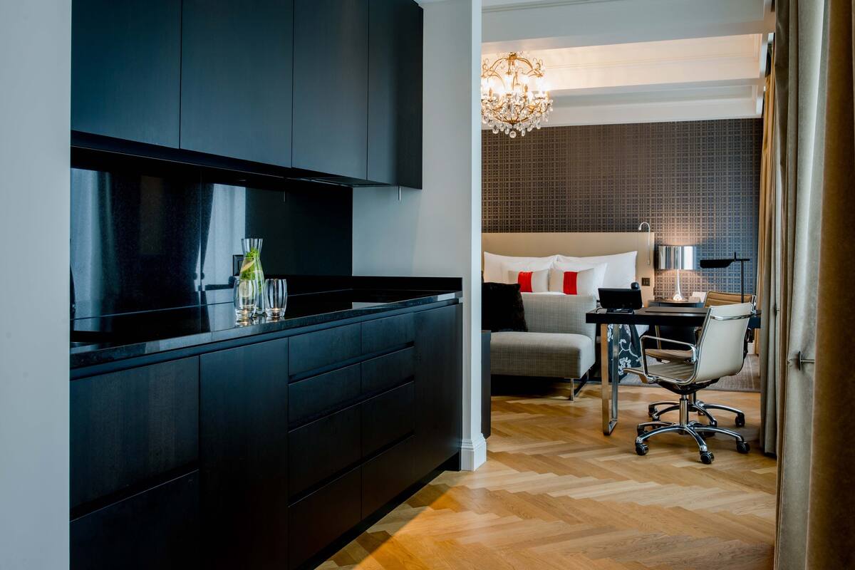 A modern hotel room with a dark wood cabinet, a bed with white and red pillows, a desk with a laptop, and a chandelier.