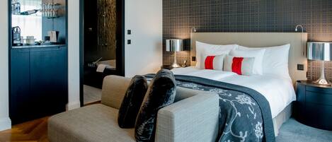 Une chambre d’hôtel moderne équipée d’un lit, d’un canapé, d’un bureau et d’une salle de bain visible à travers une porte ouverte.