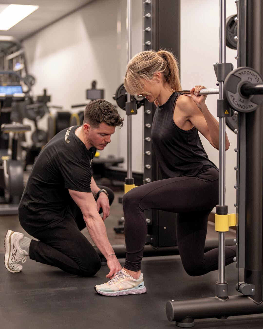 A gym with weightlifting equipment and a person assisting another with a squat exercise.