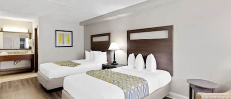 Habitación de hotel con dos camas, una mesita, una silla, una mesita de noche con lámpara y un baño a la vista.