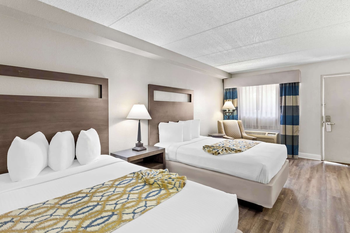 Une chambre d’hôtel avec deux lits, une table de chevet, une lampe, une chaise, une fenêtre avec des rideaux et une porte.