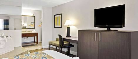 A hotel room with a bed, a desk, a television, a chair, a desk lamp, a mirror, and a bathroom with a sink and towel rack.