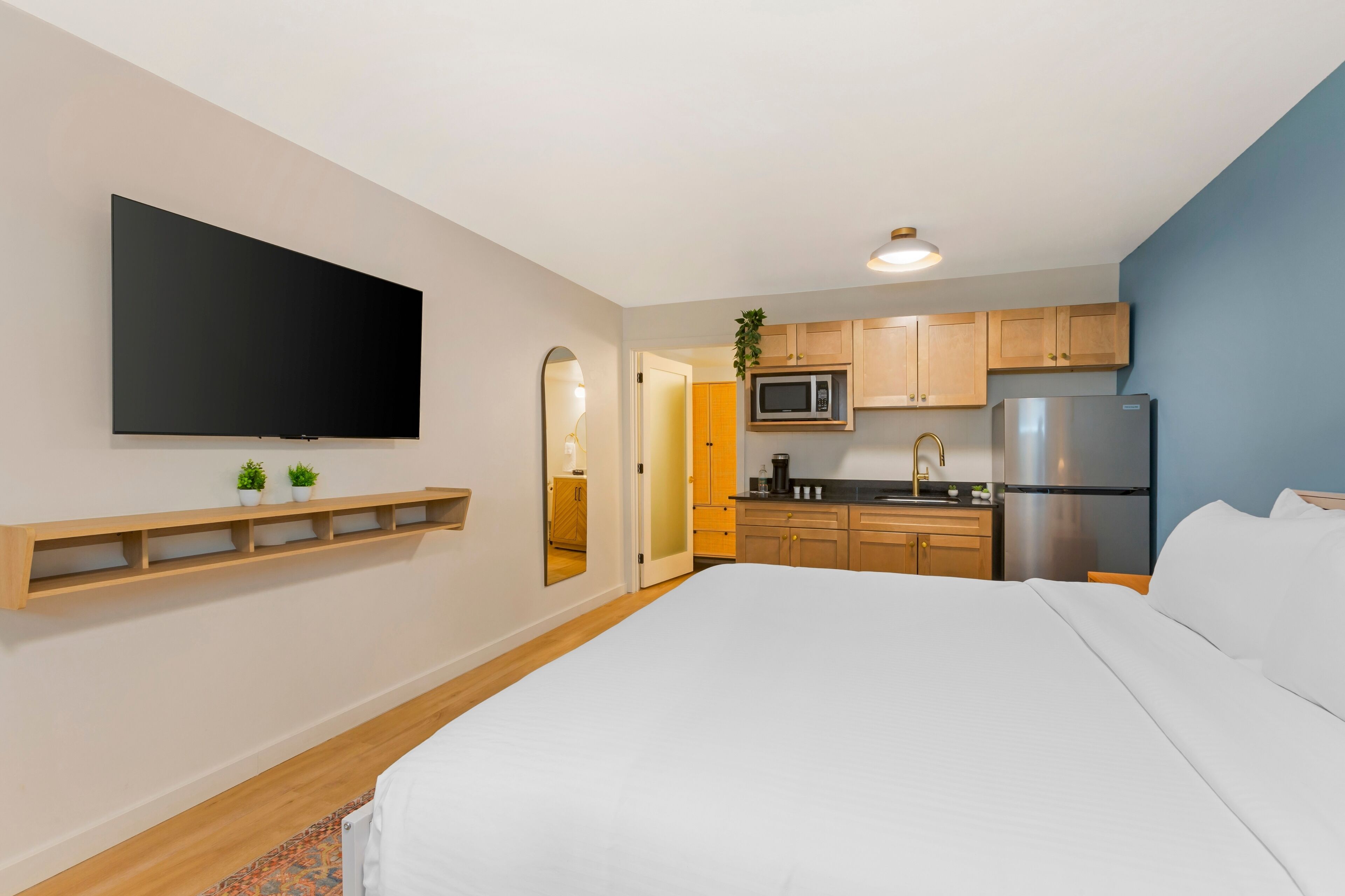 A modern hotel room with a large bed, a flat-screen TV mounted on the wall, a kitchen area with wooden cabinets and stainless steel appliances, and a small plant on the counter.