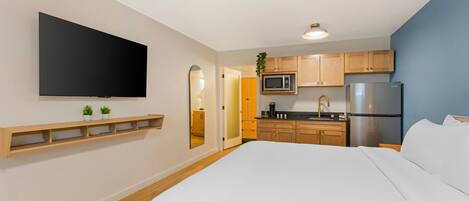 A modern hotel room with a large bed, a flat-screen TV mounted on the wall, a kitchen area with wooden cabinets and stainless steel appliances, and a small plant on the counter.