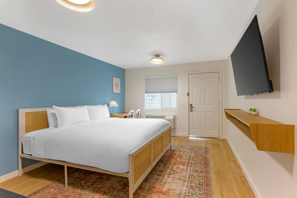 A bedroom with a bed, a bedside table with a lamp, a wall-mounted TV, and a window with blinds.