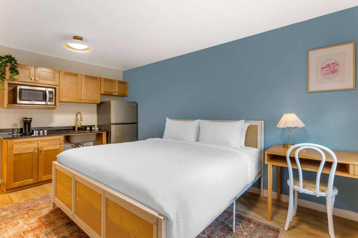 A bedroom with a bed, a desk, a chair, a microwave, a refrigerator, and a sink in a kitchenette.