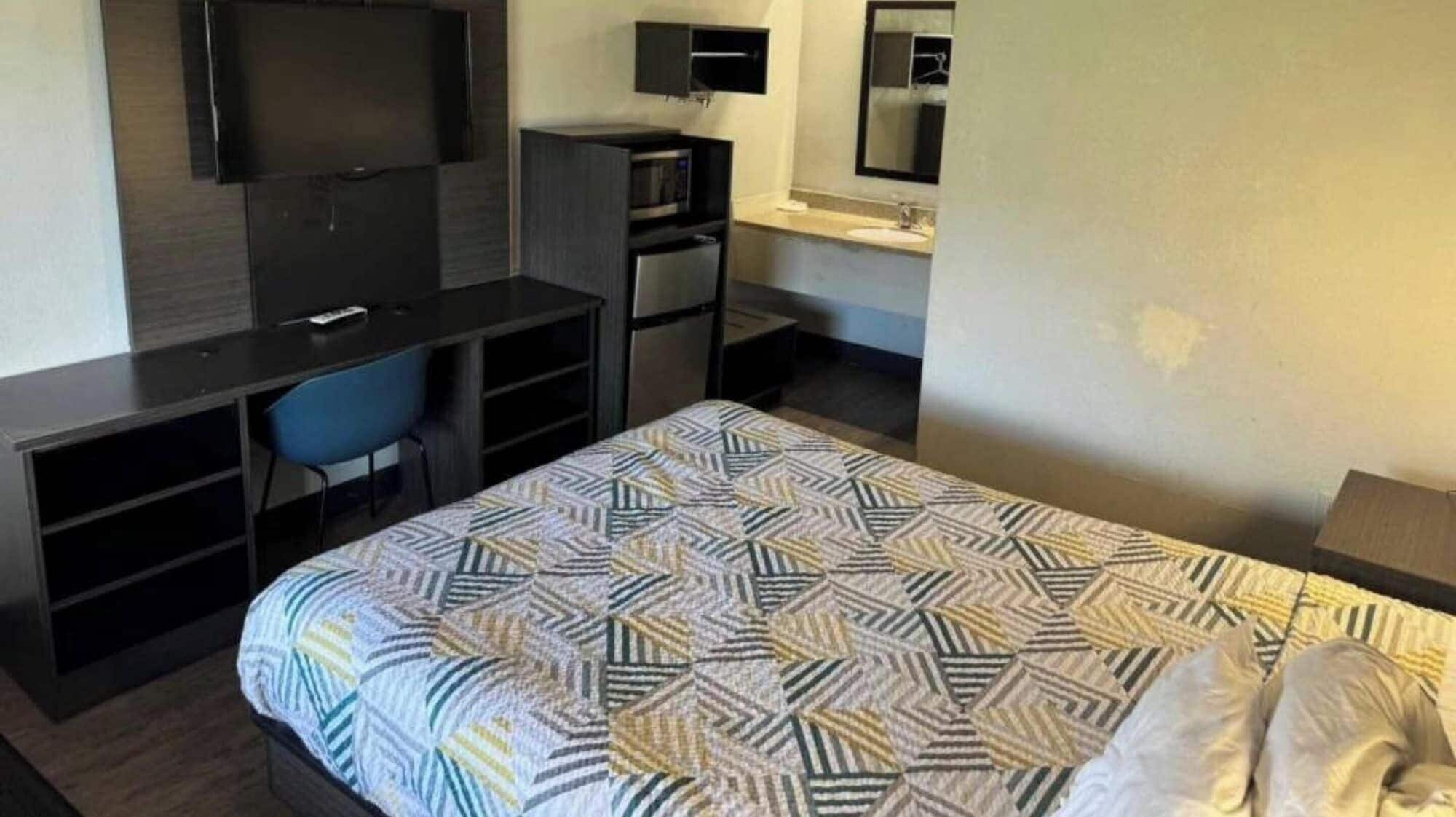 A hotel room with a bed, a desk with a chair, a TV, a microwave, and a small bathroom with a mirror.