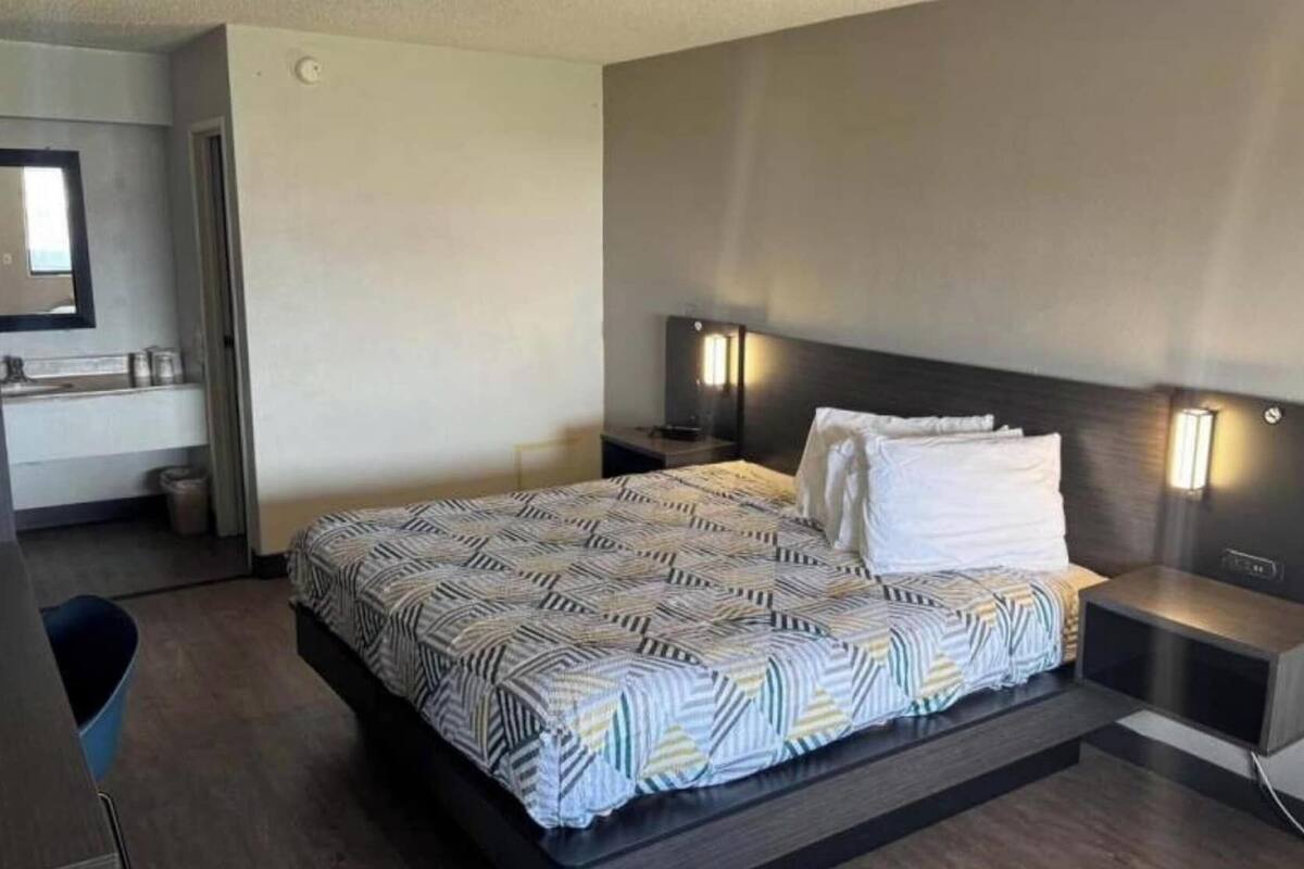 A modern hotel room with a large bed, two bedside tables with lamps, a desk with a chair, and a small kitchenette.