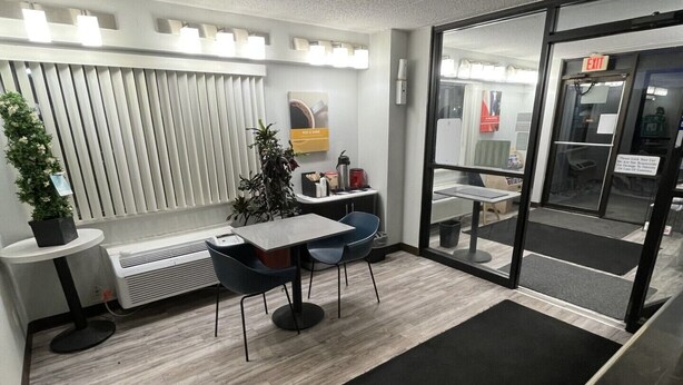 Un espacio de oficina moderno con mostrador de recepción, zona de estar con mesas y sillas, una planta en maceta y una puerta de vidrio que conduce a una salida.