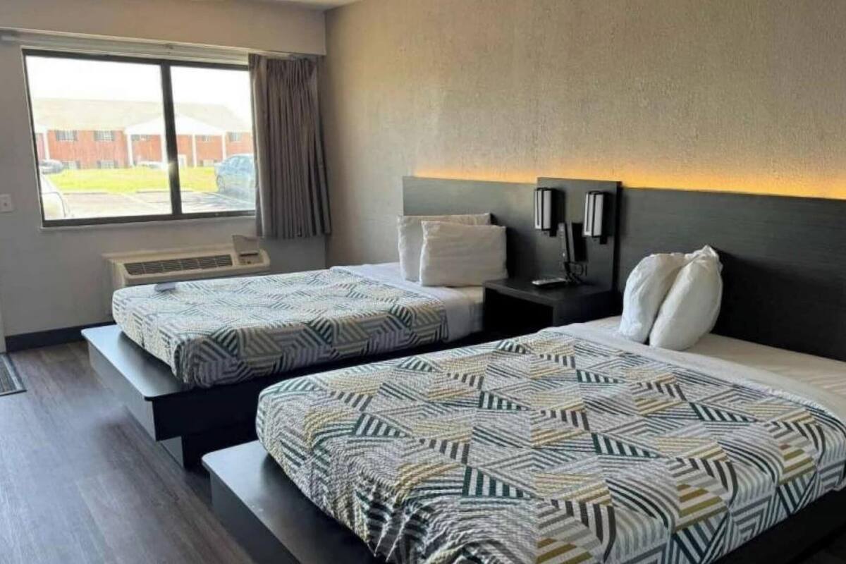 A hotel room with two beds, a window with curtains, a bedside table with a lamp, and a view of the outside.