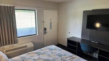 Et hotelværelse med seng, skrivebord, stol, fjernsyn og et vindue med gardiner.