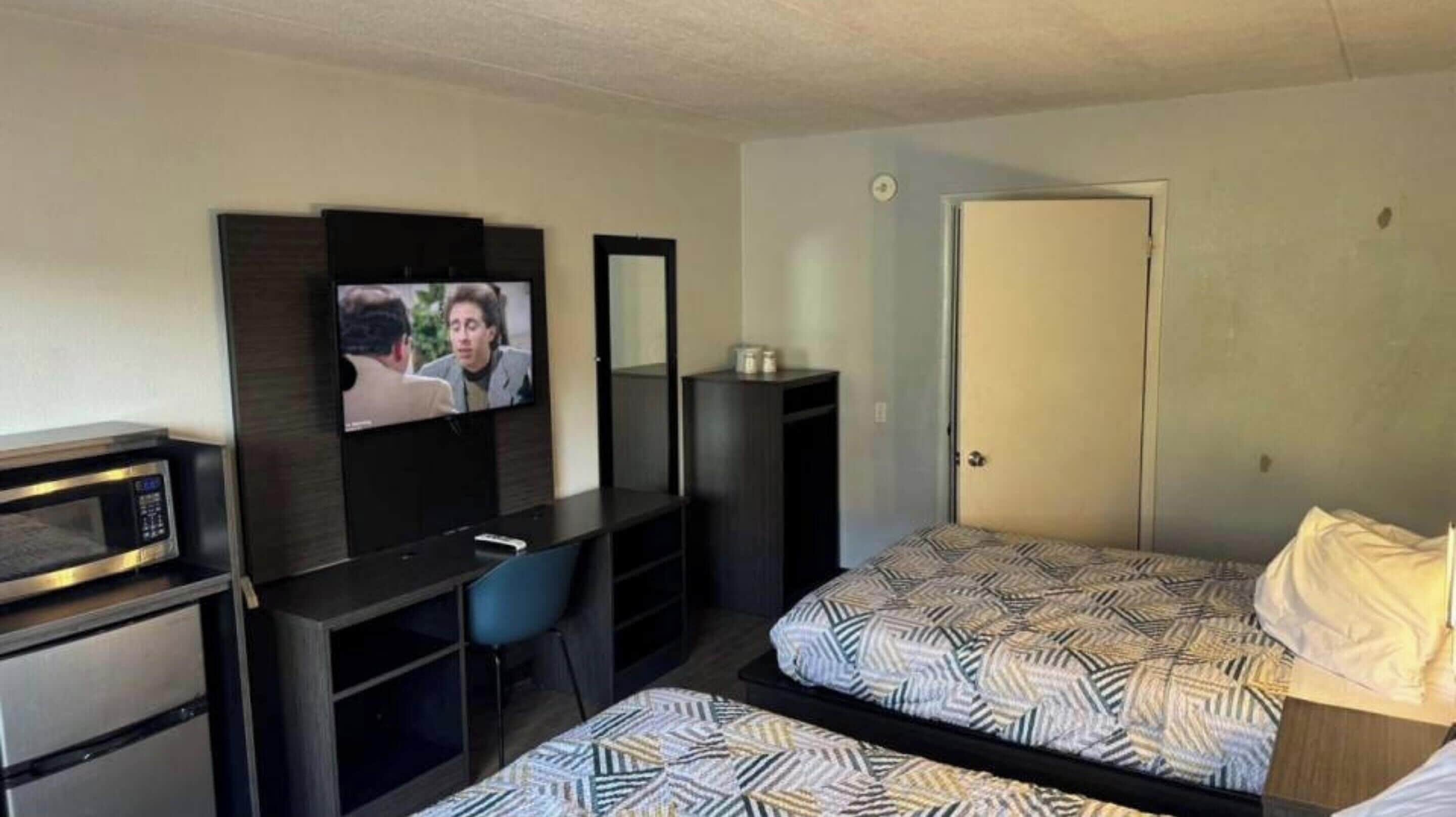 A hotel room with a bed, a desk with a computer, a TV, a microwave, and a door.