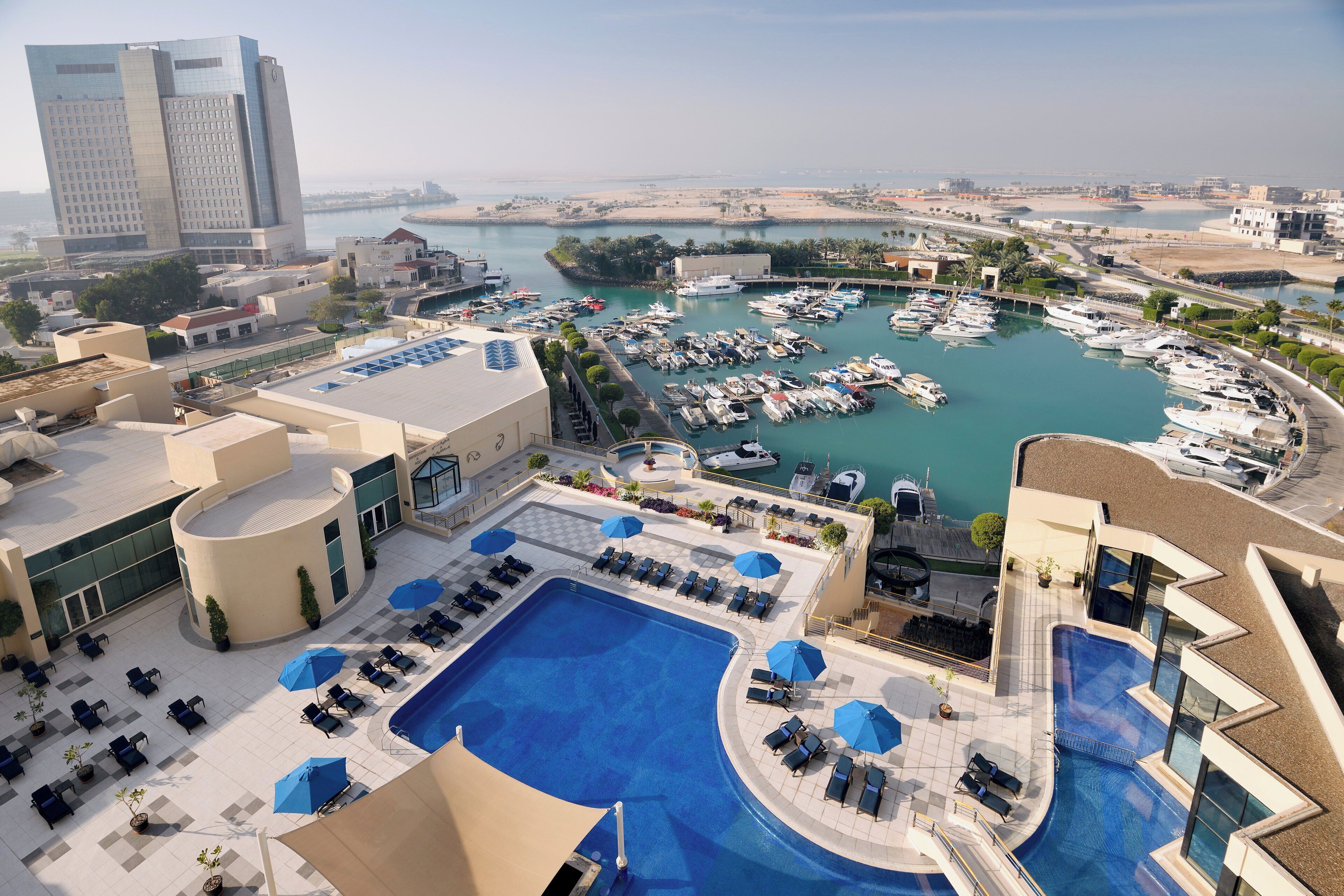 Um hotel com piscina no terraço, uma marina com barcos e vista para a costa.