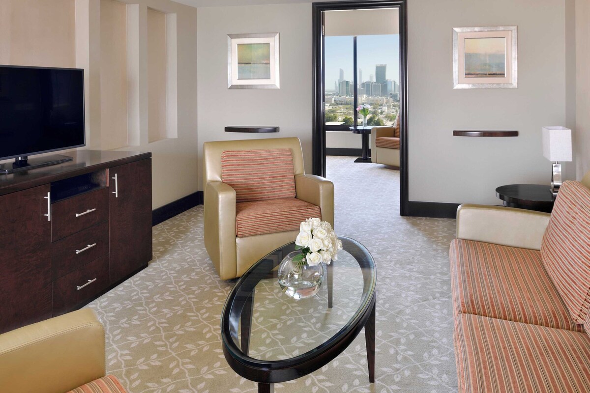 A hotel room with a TV, a sofa, a chair, a coffee table, and a view of the cityscape.