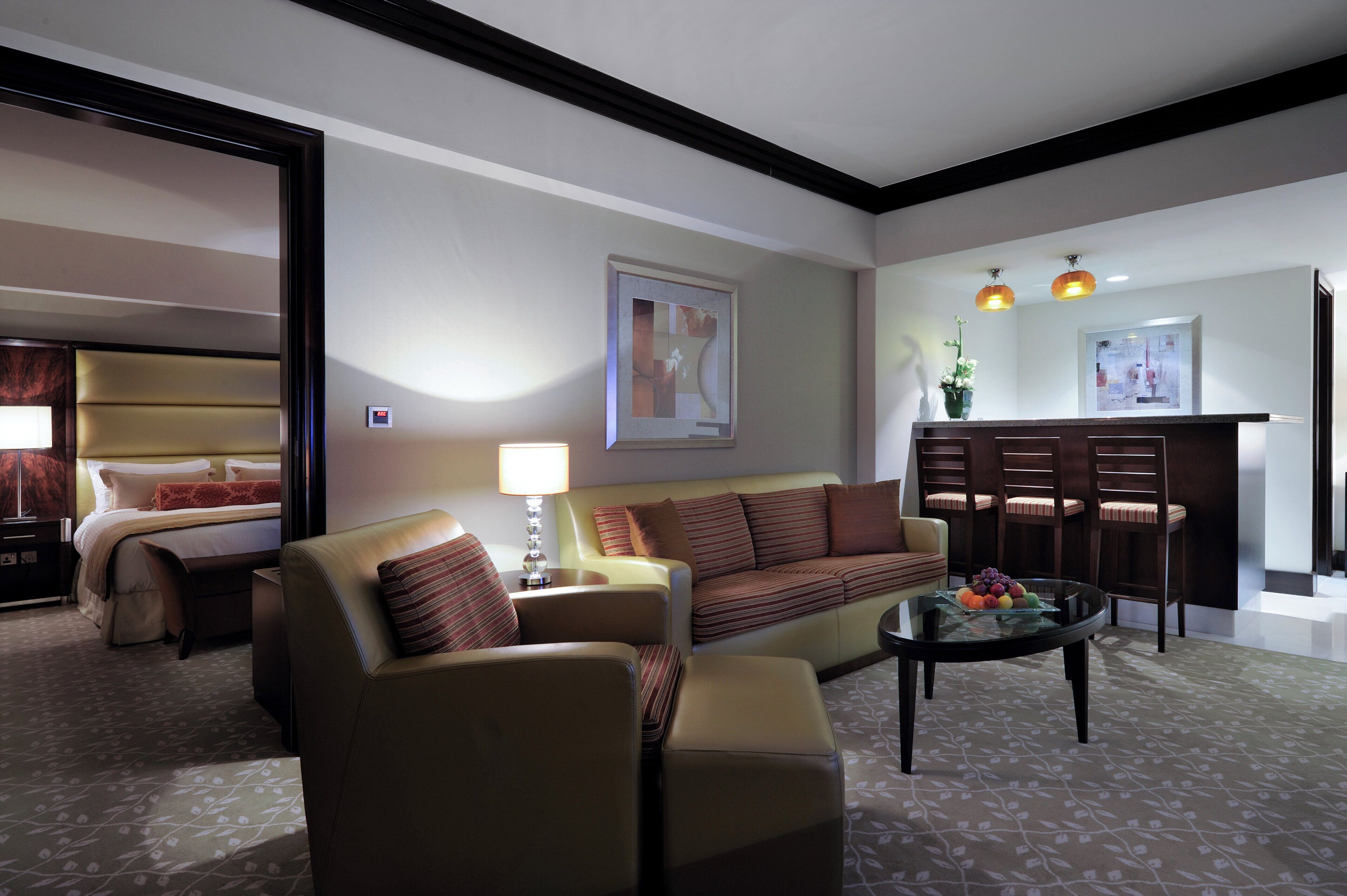 A hotel room with a sofa, armchair, a dining table with fruit, a lamp, a painting on the wall, and a bed with a headboard.