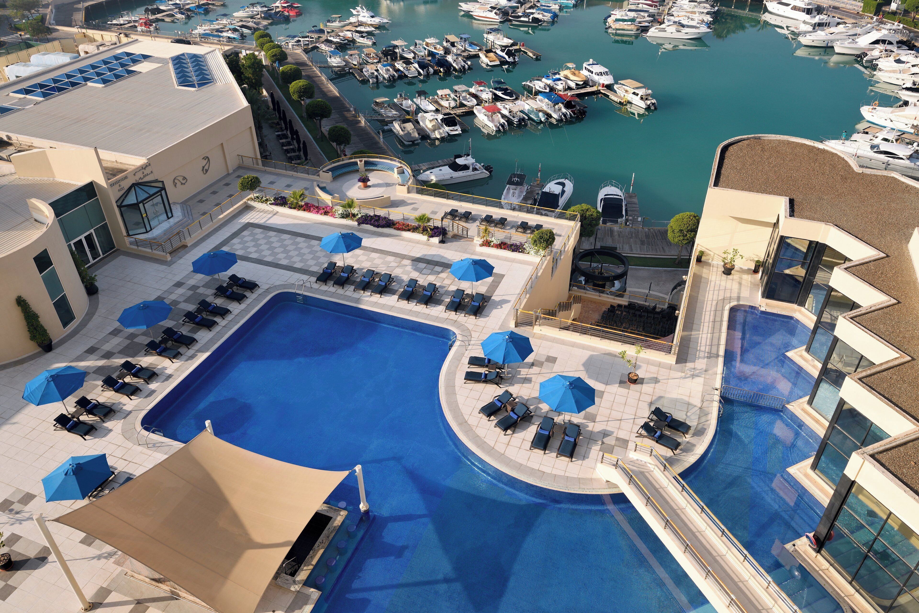 A rooftop pool area with sun loungers and umbrellas, a marina with boats, and a building with a swimming pool.