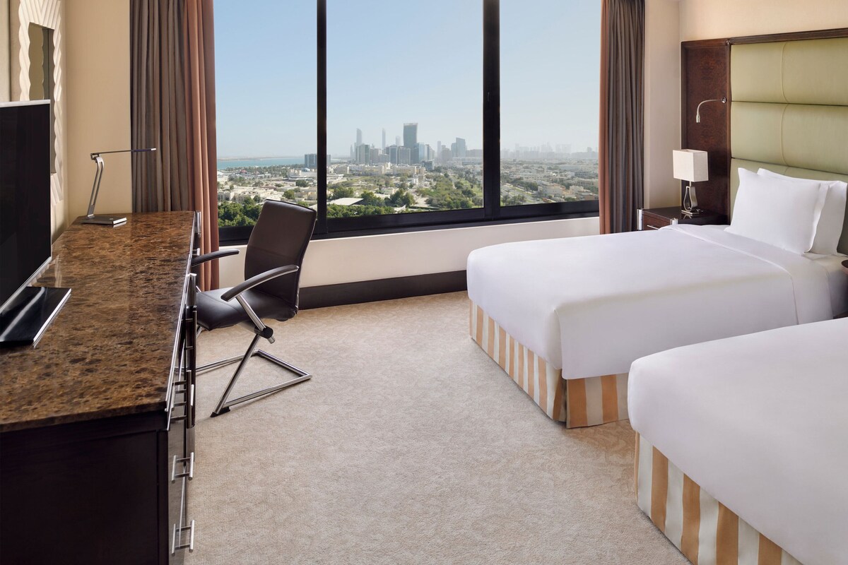 Quarto de hotel com uma mesa, duas camas, uma cadeira e vista para a paisagem urbana.