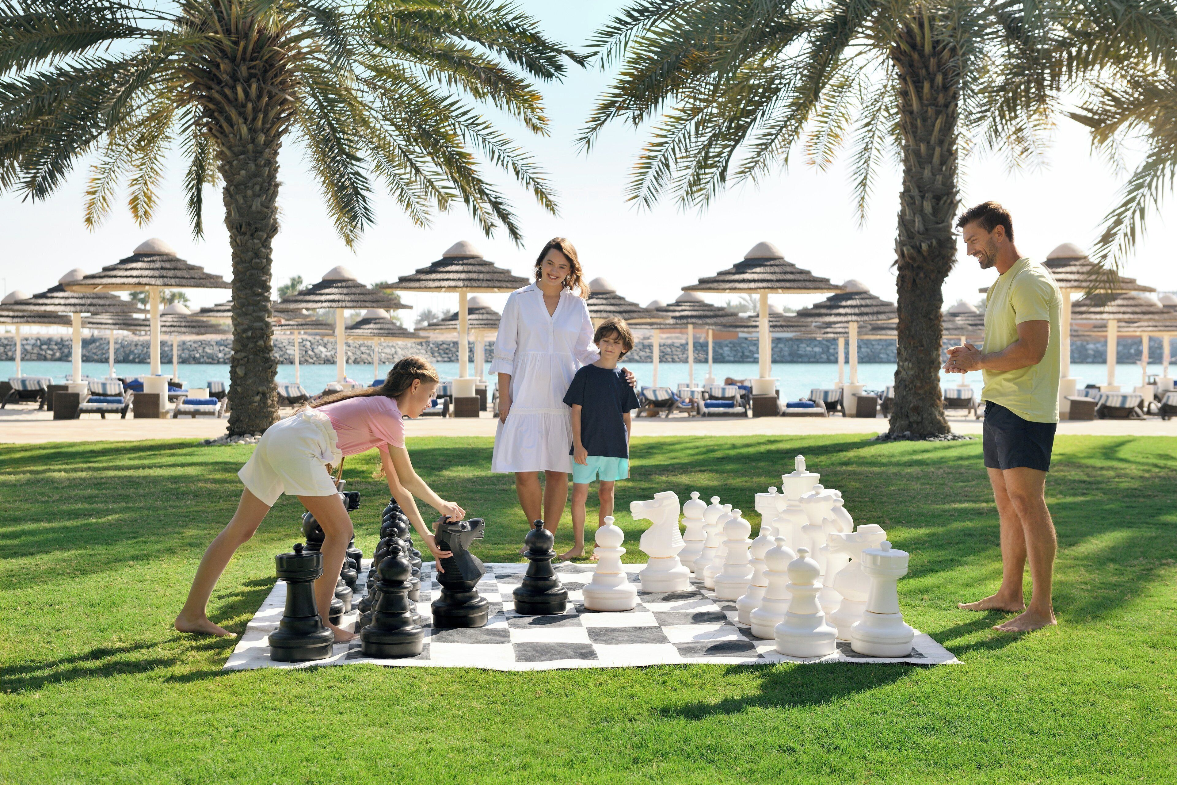 A group of people playing chess on a checkered mat in a park with palm trees and umbrellas in the background.