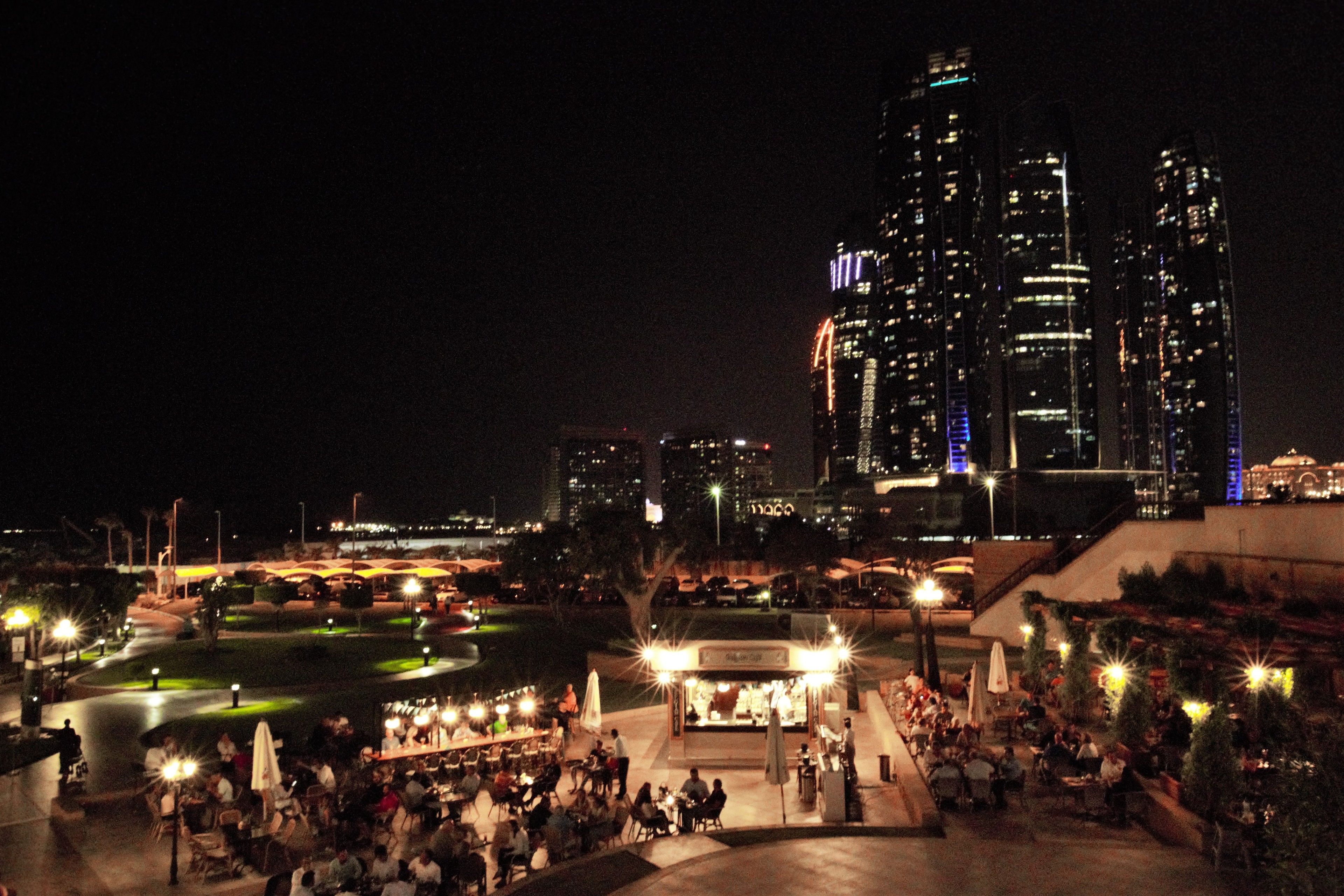 Une vue nocturne d’une ville, avec des gratte-ciel illuminés et une zone de restauration en plein air animée, avec des tables et des chaises.