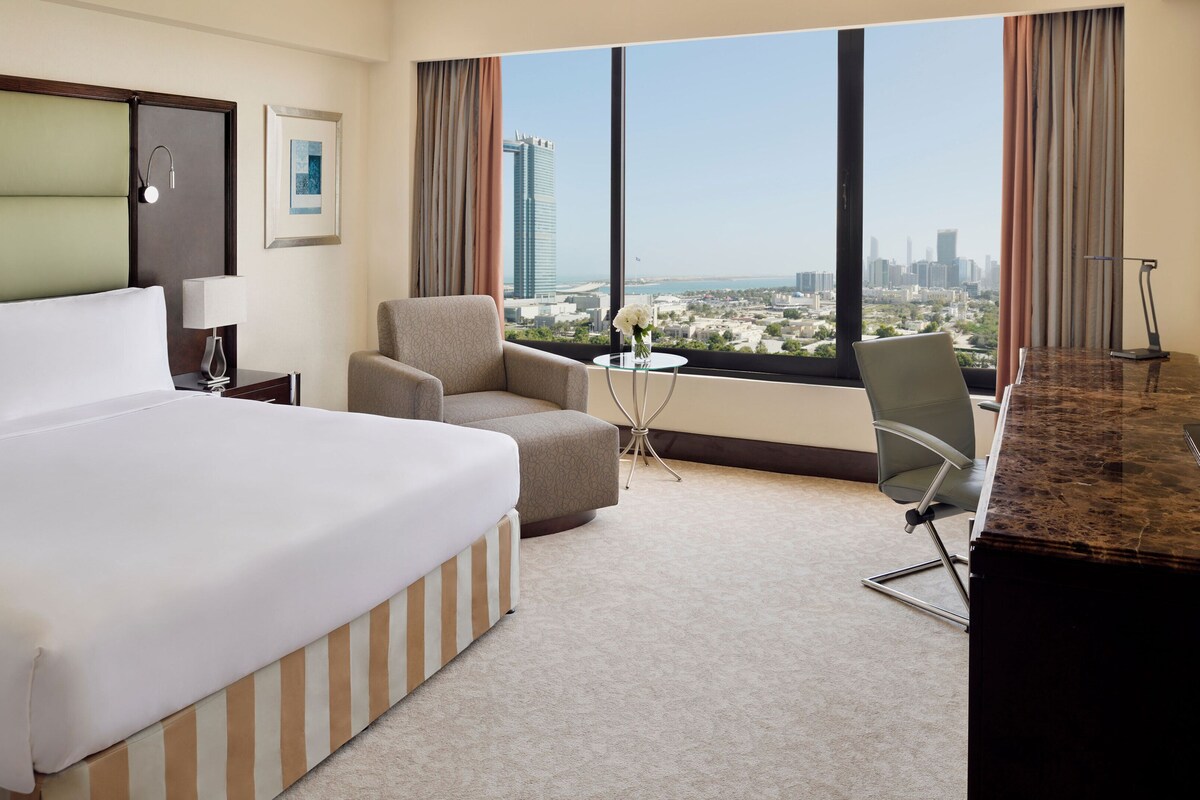 Quarto de hotel com uma cama grande, duas poltronas, uma mesinha com um vaso, televisão e vista para a paisagem urbana.