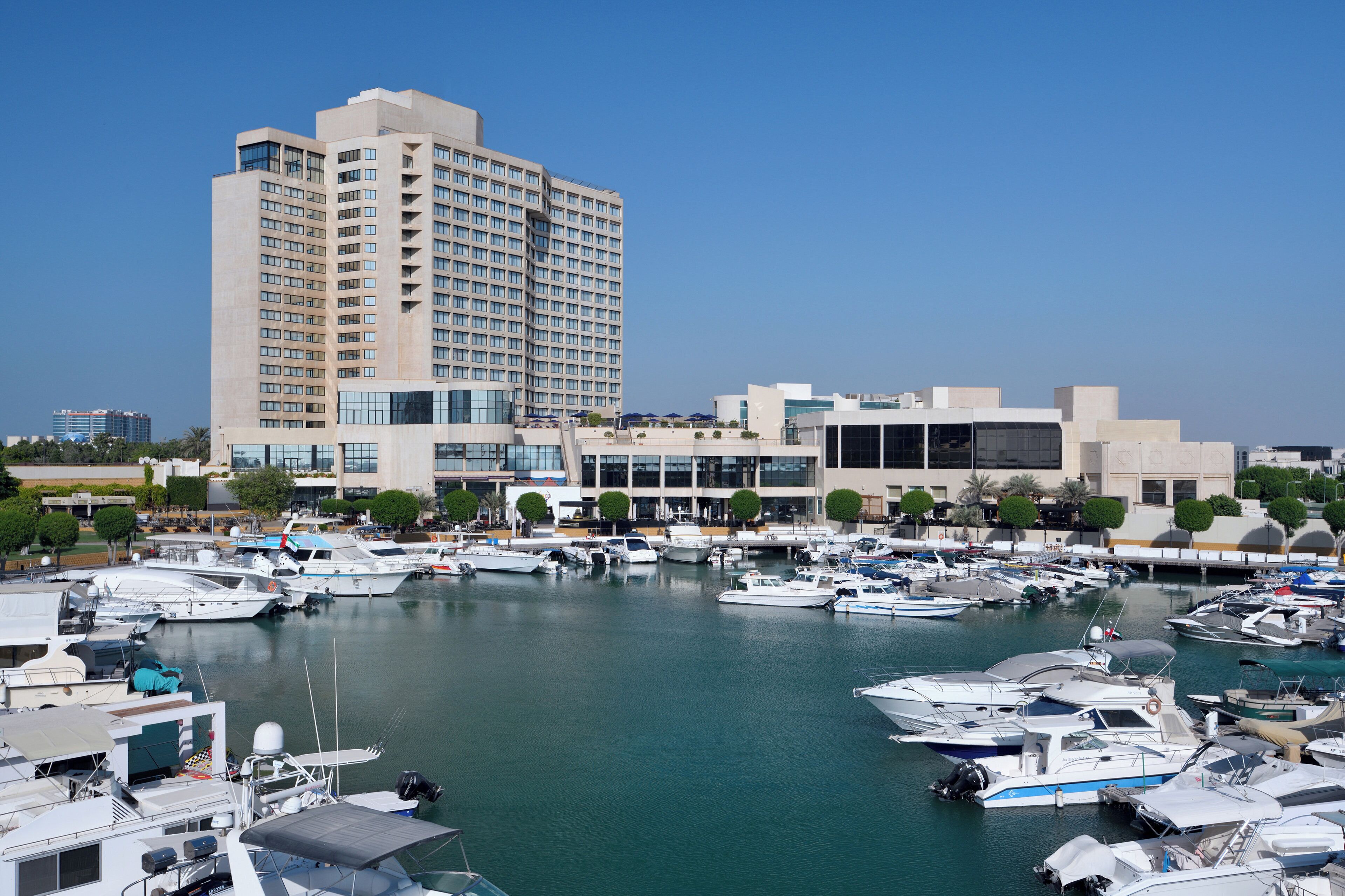 Un port de plaisance où sont amarrés de nombreux bateaux, un grand bâtiment d’hôtel et une structure moderne aux fenêtres en verre.