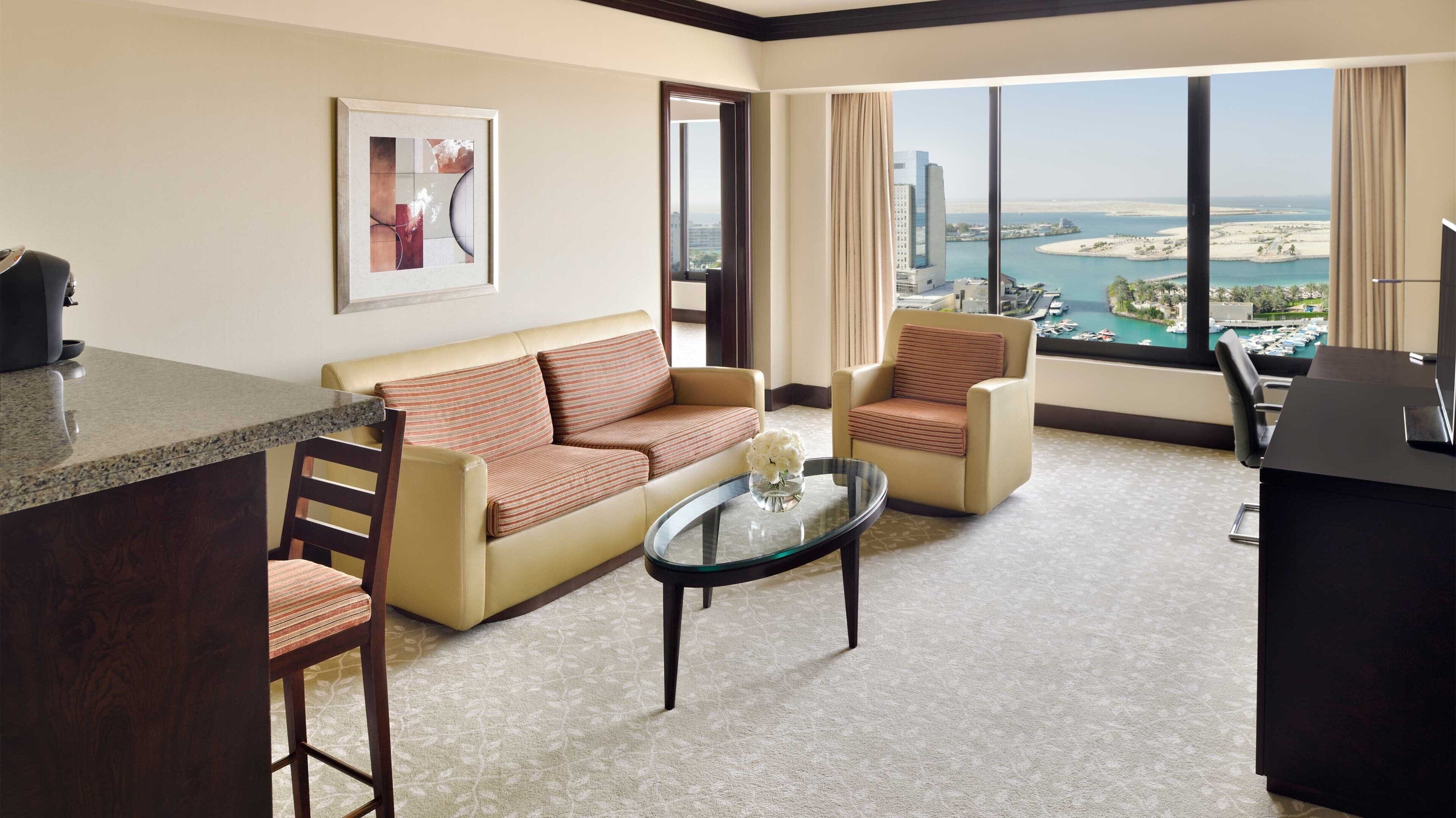 A hotel room with a sofa, armchairs, a coffee table, a desk, and a large window with a view of a city and water.