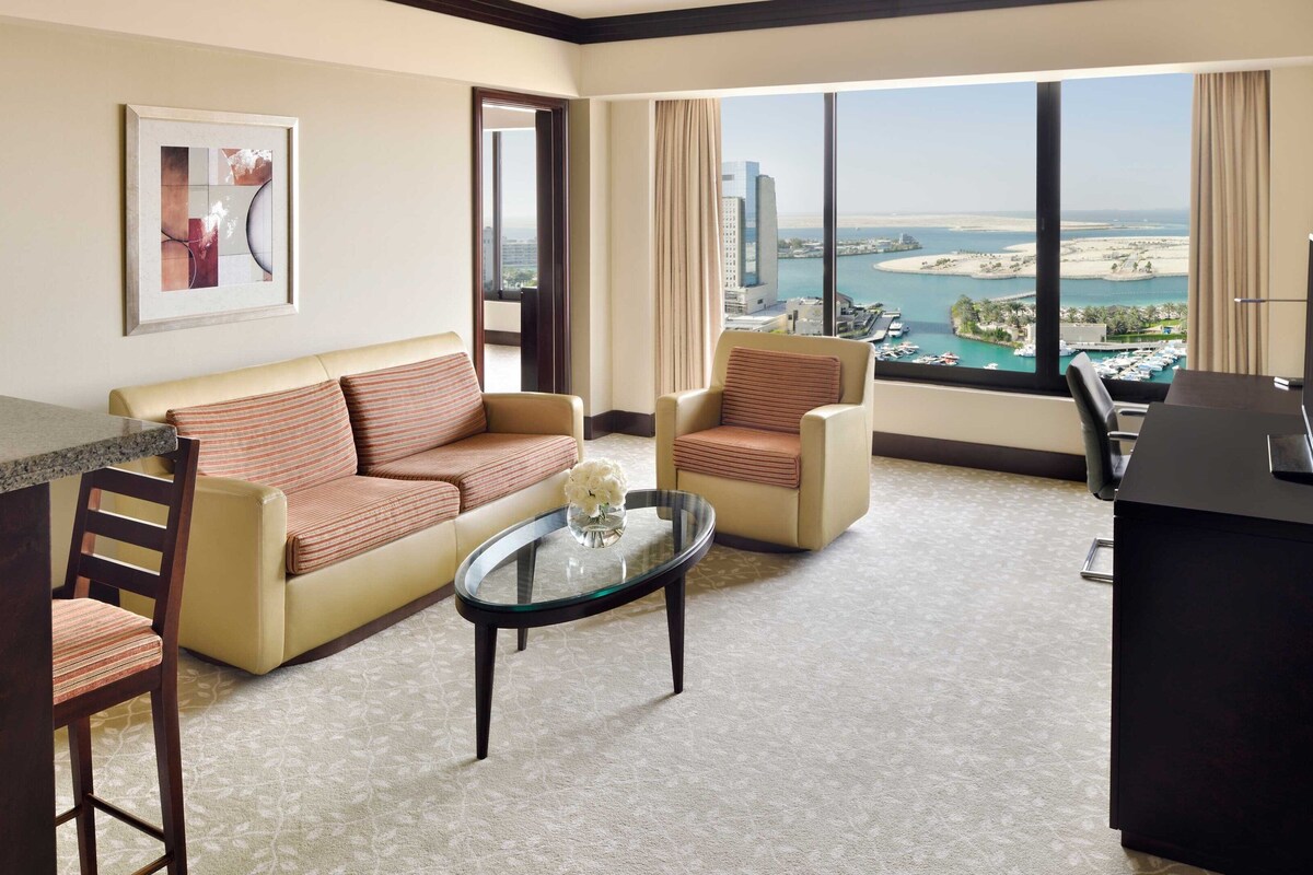 A modern hotel room with a sofa, armchairs, a coffee table, and a view of a cityscape and water.