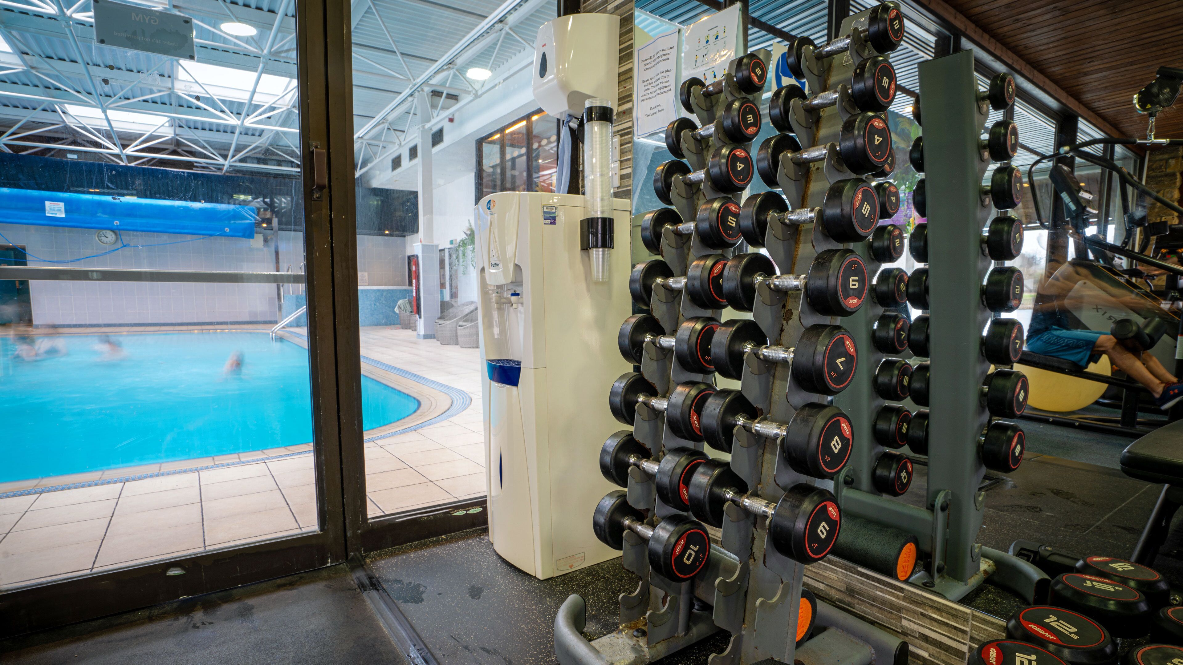 A gym with a weightlifting rack filled with dumbbells, a water pool visible through the glass doors, and a treadmill in the background.