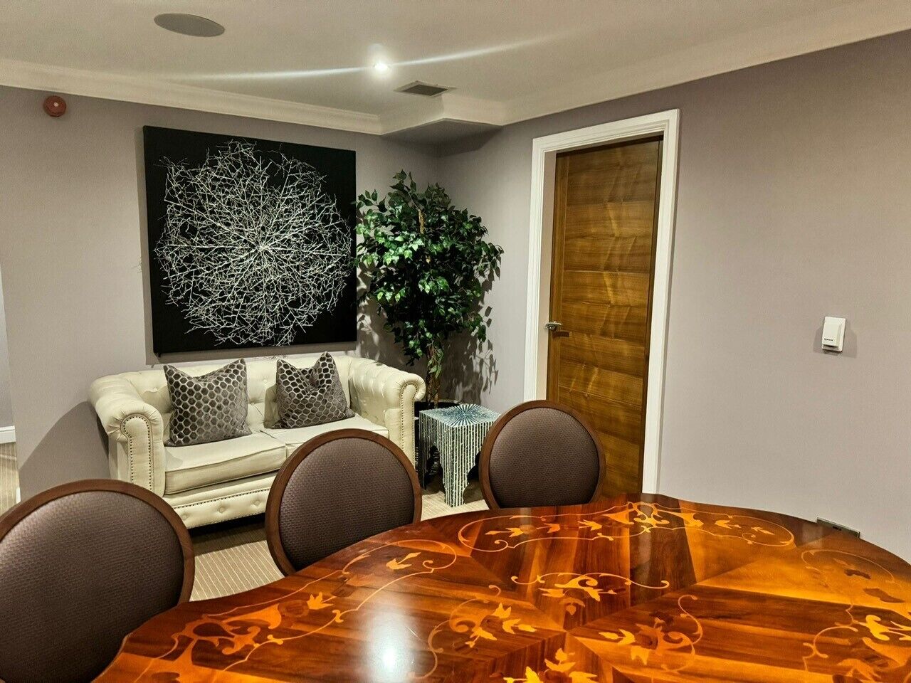 A dining area with a round wooden table, four chairs, a white sofa with patterned cushions, a large abstract painting, a potted plant, and a wooden door.