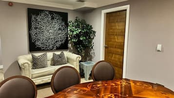 Un comedor con una mesa de madera redonda, cuatro sillas, un sofá blanco con cojines estampados, un cuadro abstracto grande, una planta en maceta y una puerta de madera.