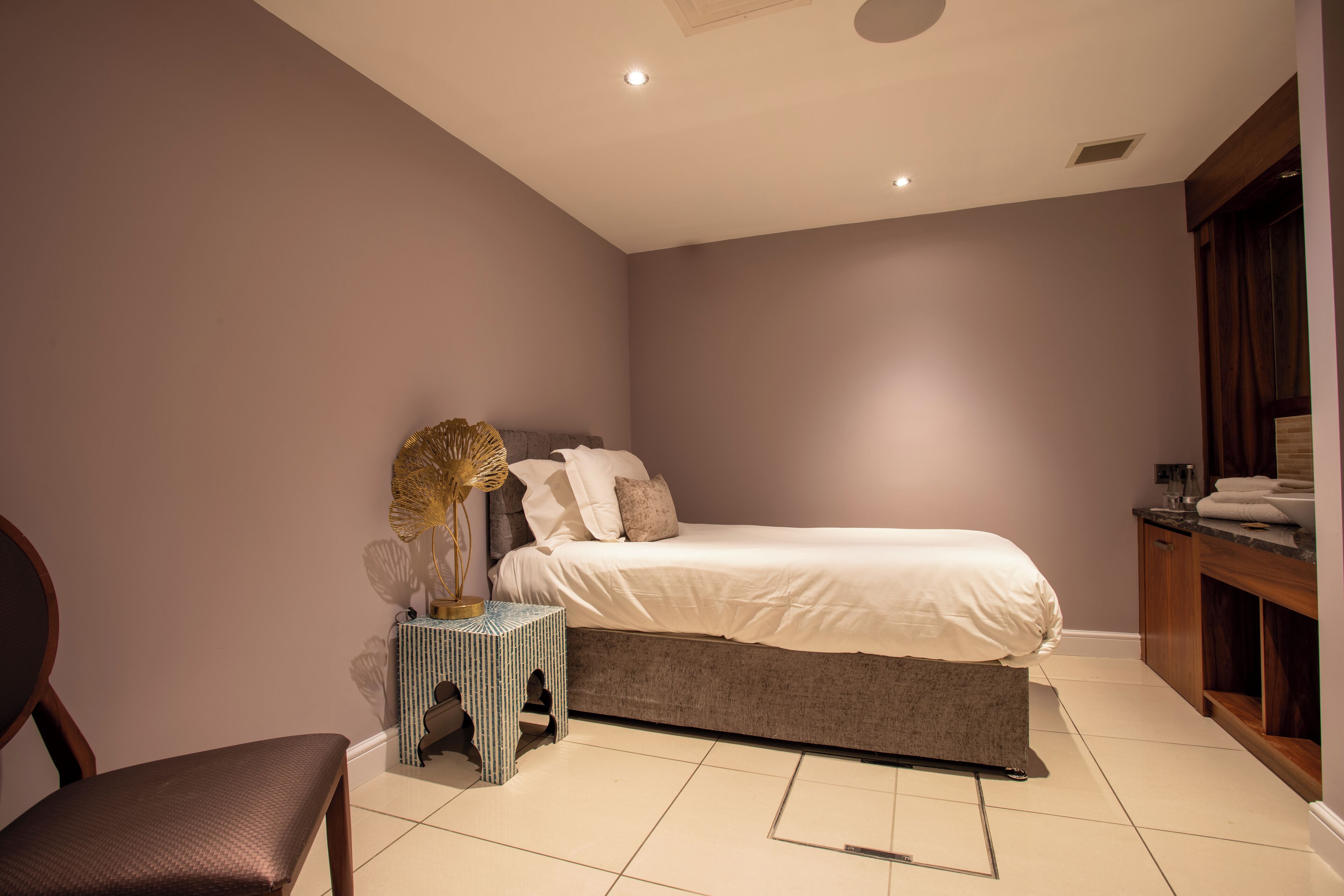 A bedroom with a bed, a chair, a small table with a lamp, and a wooden cabinet.