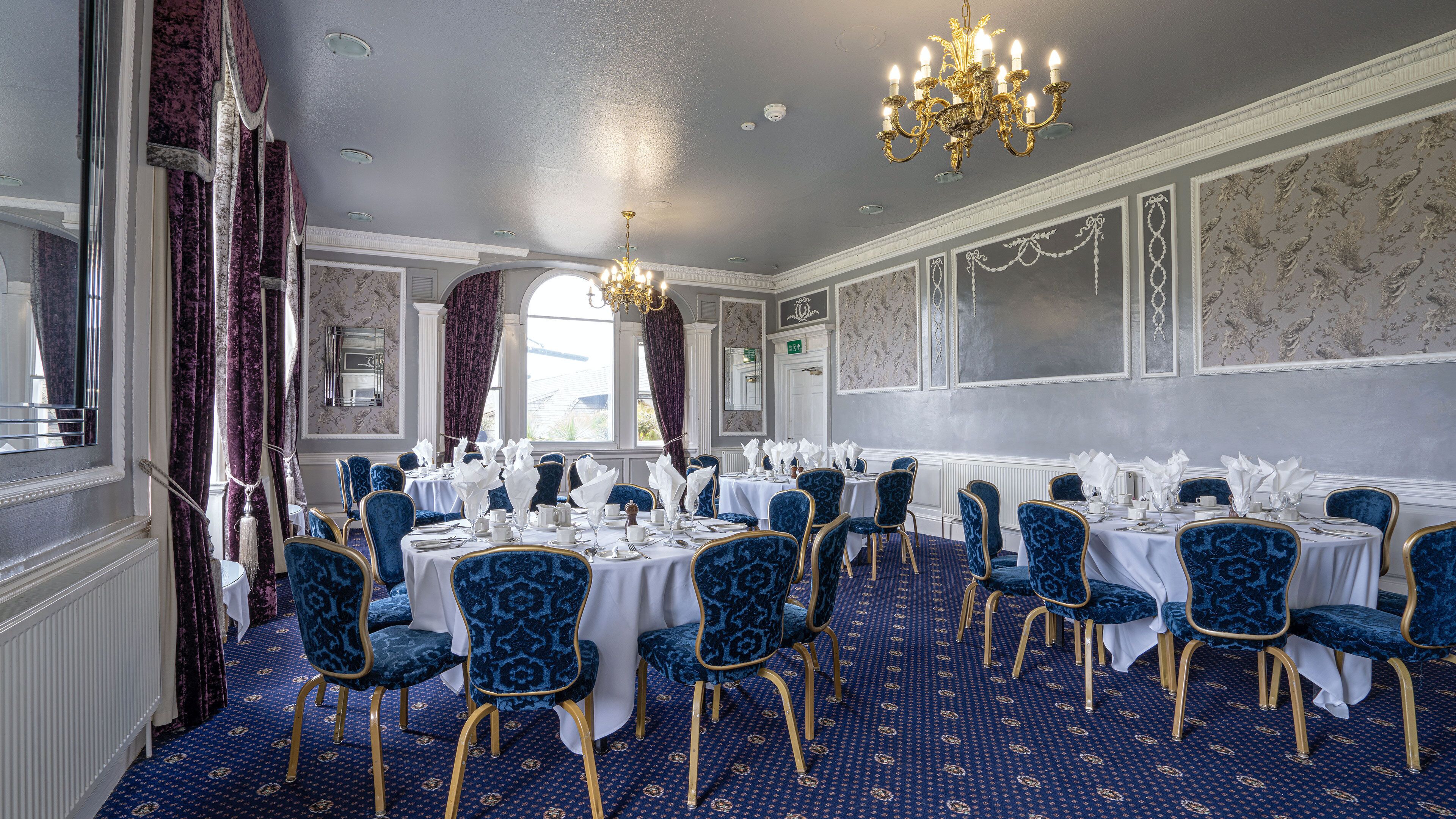 A formal dining room with round tables, blue upholstered chairs, and a chandelier.