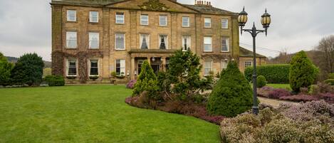 A large, historic building with a symmetrical facade, multiple windows, and a well-maintained lawn with various shrubs and trees.