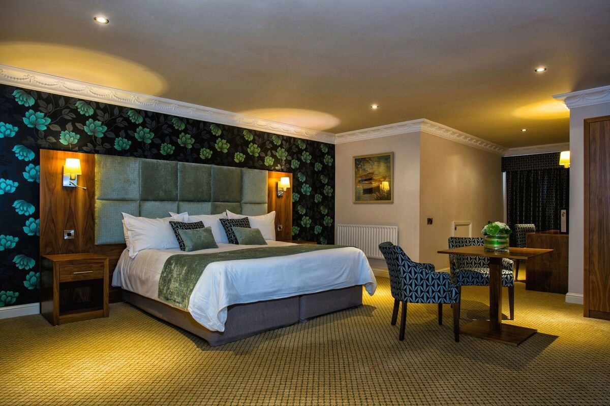Quarto de hotel com uma cama grande, mesa de cabeceira, cadeira, mesinha e radiador.
