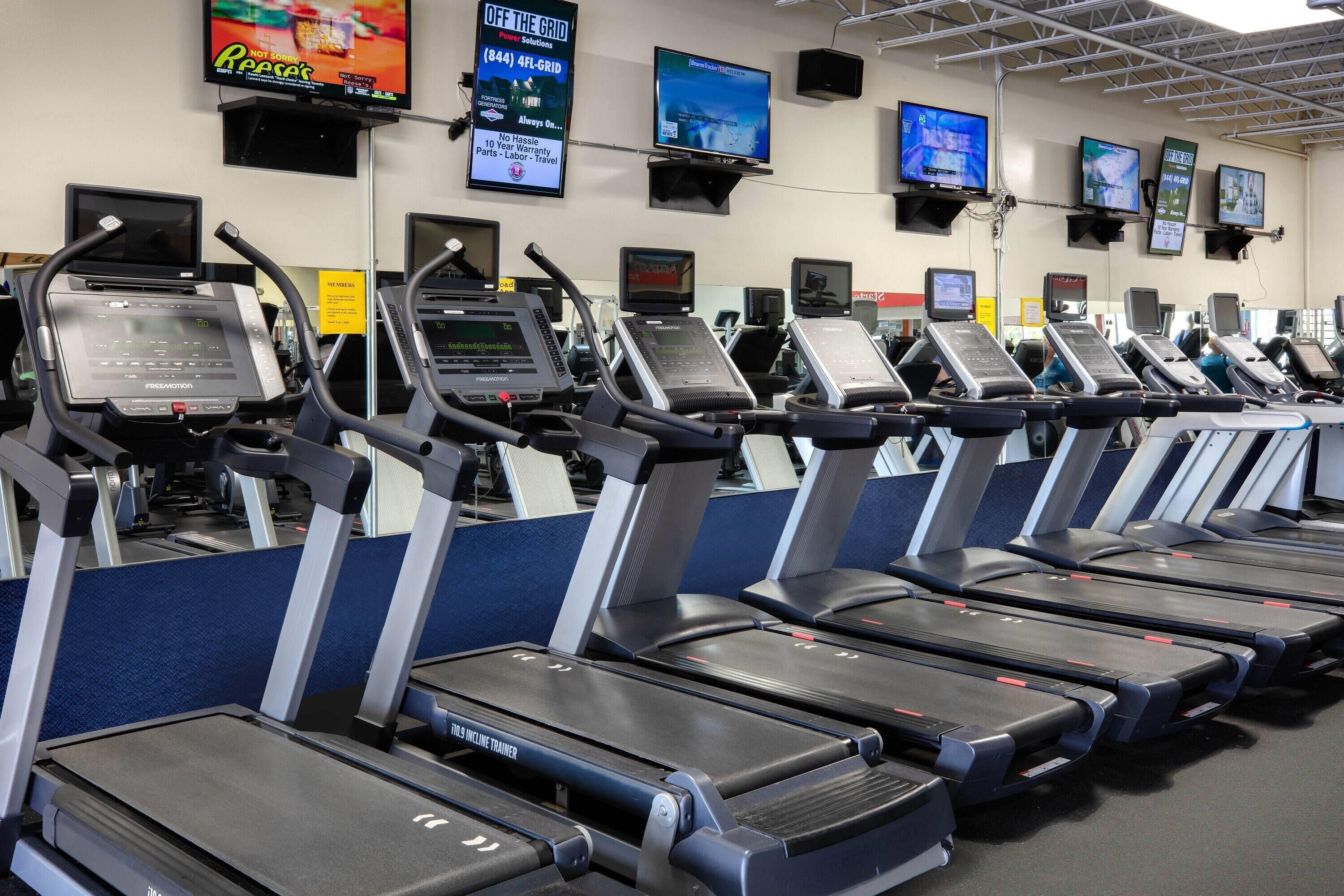 A row of treadmills in a gym with multiple screens displaying various content.