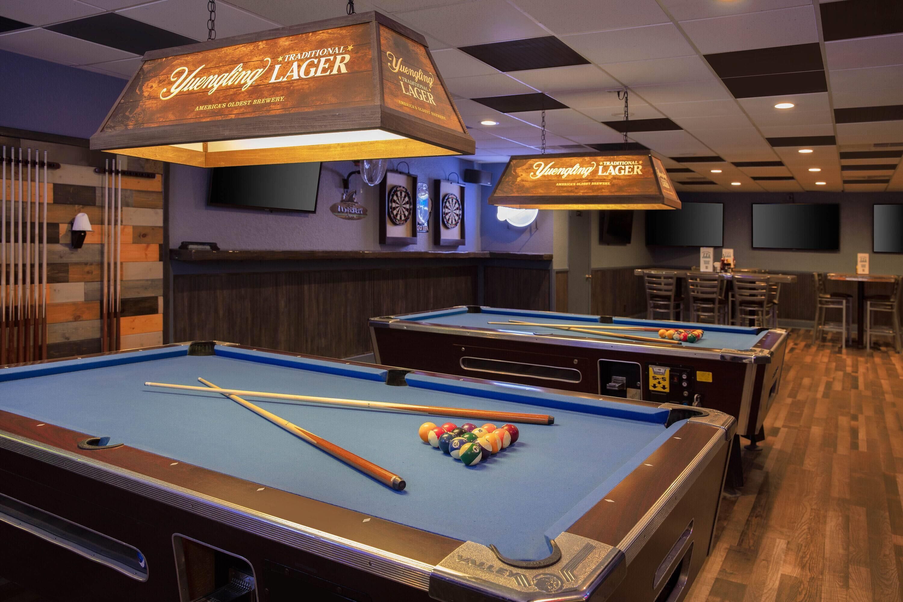 Une table de billard avec des queues et des billes, un jeu de fléchettes et un coin bar avec une enseigne indiquant « Yuengling Lager ».