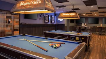 A pool table with cues and balls, a dartboard, and a bar area with a sign reading "Yuengling Lager".