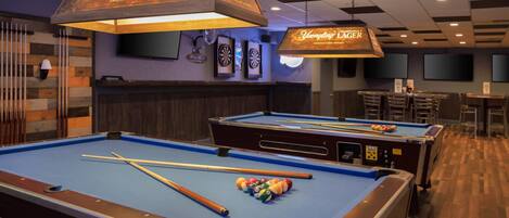 Une table de billard avec des queues et des billes, un jeu de fléchettes et un coin bar avec une enseigne indiquant « Yuengling Lager ».